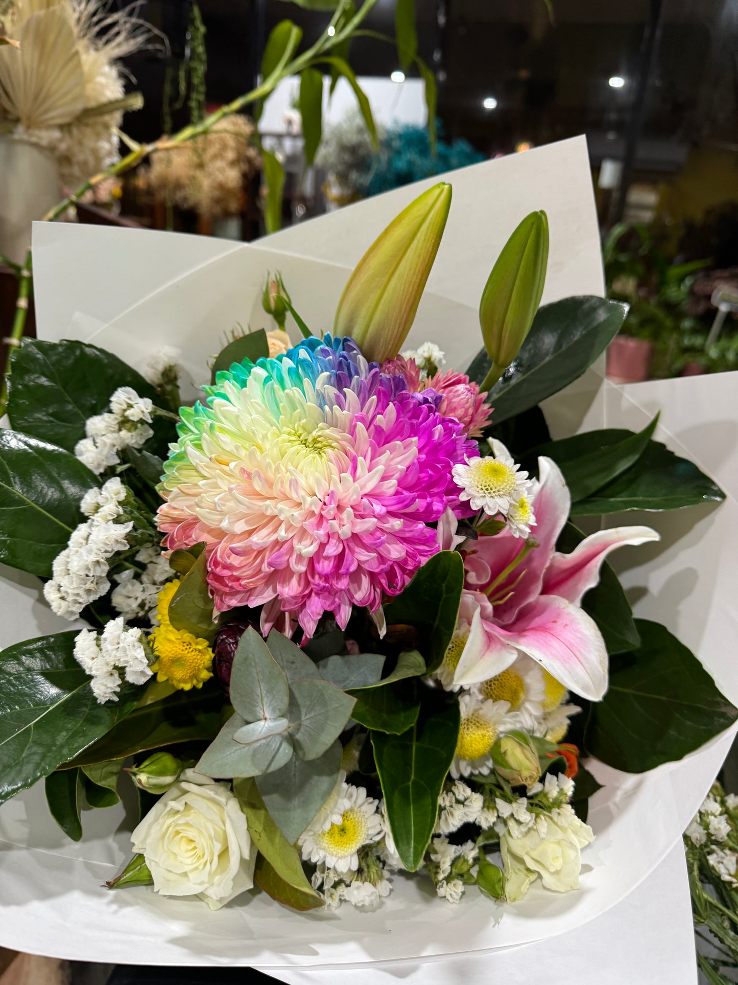 Bouquet of flowers with a colourful flower in the centre, arranged with Oriental lilies,
statice and daisies with beautiful dark foliage framing the bouquet wrapped in white paper.